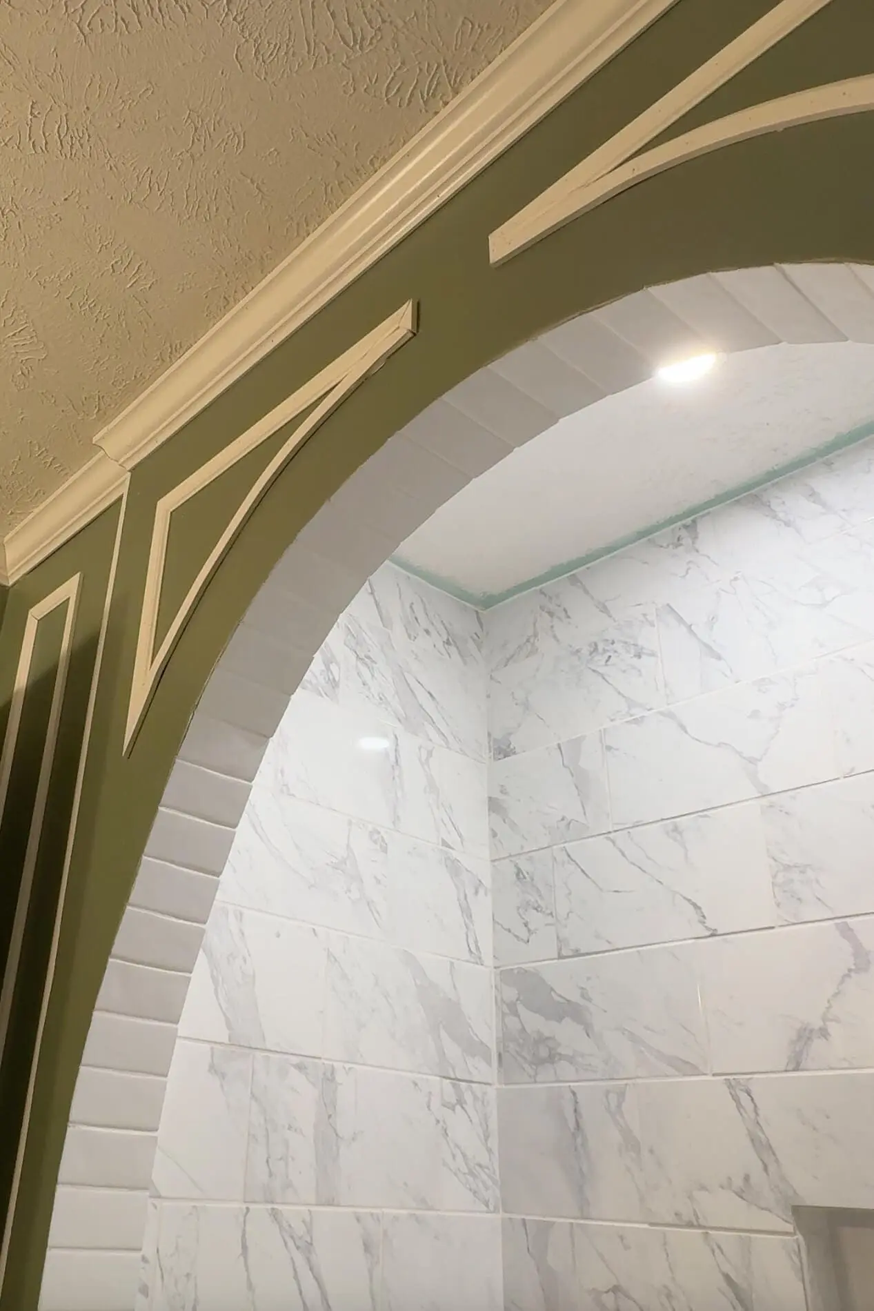 Elegant shower archway with green walls featuring white curved trim. The shower interior showcases glossy white marble-style tiles with a recessed shelf and a modern gold rain showerhead, highlighted by recessed lighting above.